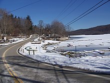 A road straddling the bank of a frozen lake. The surrounding ground is covered by thin layer of snow. Powerlines are visible overhead