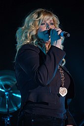 A woman with long blonde hair wearing dark clothing and singing into a microphone