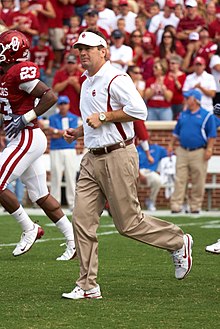 A man jogs onto an American football field wearing a visor, polo and khaki pants, with several football players and fans in the background.