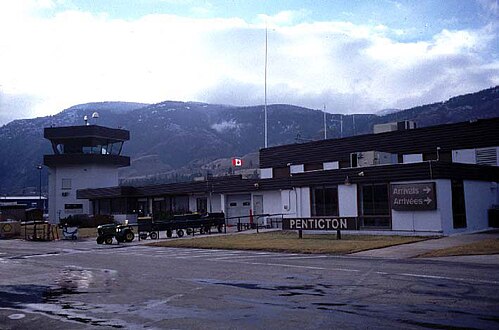 The main terminal building of an airport, with a "Penticton" sign near the entrance, as well as a "Arrivals" sign point to the right, with the same word wrote in French below it.