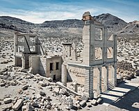 Aerial view of the same three-story white masonry bank building in 2022, roofless with large sections of its walls missing. Masonry rubble lies about the building, which has openings for doors and windows but no glass or wood. The collapsed walls of another, smaller ruin are to the right. Barren hills are visible in the distance.