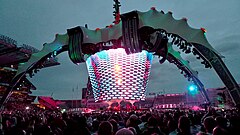 A concert stage; four large legs curve up above the stage and hold a video screen which is extended down to the band. The legs are lit up in green. The video screen has multi-coloured lights flashing on it. The audience surrounds the stage on all sides.