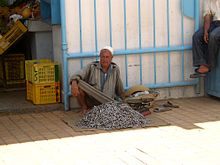 Vendedor de pescado sentado sobre la acera y apoyado en un muro; la mercancía está puesta a sus pies.