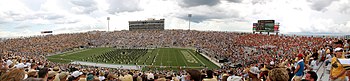 A football stadium from the upper portion of the seats with several rows of people dressed in black on the field.