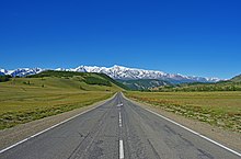 Photographie depuis le centre de la route. Le paysage est verdoyant, avec en arrière-plan des cimes enneigées, dans la steppe de Kouraï.