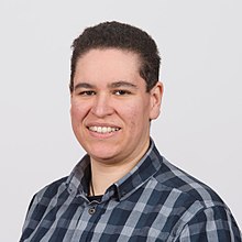 Head and shoulders image of a white person with short dark hair looking at the camera wearing a blue and white check shirt