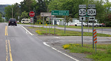 Two roads meeting at a very oblique angle with a large stop sign at the junction. Signs at the right indicate that traffic going straight ahead is following Route 30 southbound, with a right turn indicated for Route 30A northbound. Between the two signs a white-on-green sign says that it is three miles to Schoharie straight ahead and two miles to Central Bridge on the left. Beyond the intersection are some trees, trailer homes and parked cars