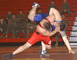 Two men wrestling in a gymnasium, watched by a group of uniformed soldiers