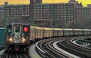 A 1 train, composed here of R62A cars is seen above ground entering the 207th Street station. The front of the train contains two white lights providing slight illumination, two windows, a door, and the Symbol for the 1 line on the left window.