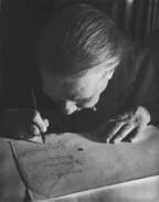 Photographie en noir et blanc du haut du crâne dégarni d'un homme en train d'écrire, fortement penché sur sa feuille à cause de sa vue faible.