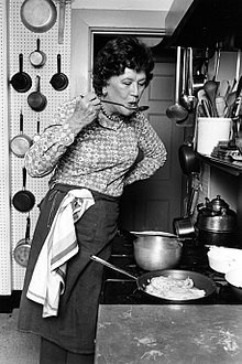 Julia Child in front of a stove tasting food on a spoon
