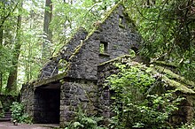 Ruins of a moss-draped stone building rest near a path in a thick forest.