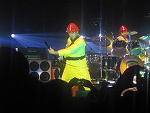 In the middle of a stage, a man wearing a yellow suit and a red hat plays a guitar. In the background is a similarly dressed man playing drums. A crowd is visible in the foreground.