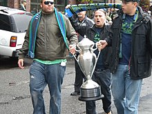 Two men carry a bright silver trophy with another man walking behind holding a green and blue scarf overhead.