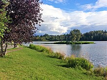 Artificial lagoon with a small island in the center. The island and bank leading down to the lagoon are grassy and have a few ornamental trees. Along the leftside there is a paved trail. On the opposite side of the lark are many trees, behind which are a few mountains.