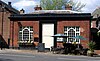 A symmetrical single-storey brick building with a central white tablet almost the height of the building, windows with complex tracery on each side, and, in the front, picnic-type tables and a menu board.