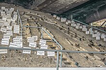 Talud sobre el cual se ve una silueta humana gigante, rodeada de carteles blancos con nombres de personas.