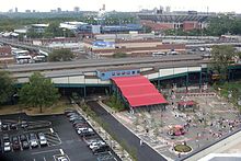 The entrance/exit stairs for the Mets-Willets Point station as viewed from Citi Field. The stairs are extremely wide and are covered by a red canopy, which leads from a plaza in the foreground to the station's mezzanine in the background. The Long Island Rail Road station is at rear left and the USTA National Tennis Center is at rear right.