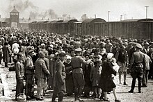 Large number of people standing beside a railway siding with the camp gate in the background