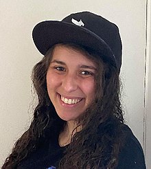 A woman with black hair wears a black sports jersey and baseball cap