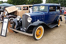 A dark green 1920s-style car with beige/cream trim facing forward angled to the picture's right in on a brick-paved lot surrounded by other cars