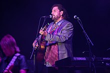 Un homme avec une légère barbe et des cheveux longs bruns, de profil, jouant de la guitare et chantant dans un micro.