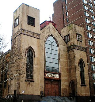 The front and part of the side of a three-story building is visible. The side is mostly hidden by the photographic angle and by a leafless tree. The front shows two rectangular towers, one on each side of a recessed bay. All are clad in tan stucco, which is stained in places. The towers have pointed arched windows on the bottom and square ones on top. The bay has four wooden doors at the bottom and a sign with Hebrew writing on top of them, surmounted by large arched, multi-paned window. Atop the roof of the bay is a small metal Star of David. To the right of the building is a much taller brown rectangular apartment building.