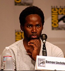 A black man wearing a white shirt. A panel with the logo for the San Diego Comic-Con is seen behind him. In front of him are a water bottle, a plastic cup, a microphone and a paper reading "Damon Lindelof".