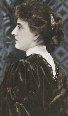 A white woman in profile. She is wearing a high-necked white blouse and a dark over dress with puffy sleeves. Her hair is back and caught in a visible hair ornament behind her head.