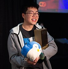 Yu smiling while holding up a golden J-Piece trophy and a blue penguin squishmallow