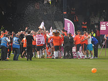 A crowd of men, some wearing grey suits and some wearing white shirts, navy shorts and white socks, celebrate raucously on a podium. An open bottle of champagne is visible in front of them, spiralling through the air as if somebody has thrown it