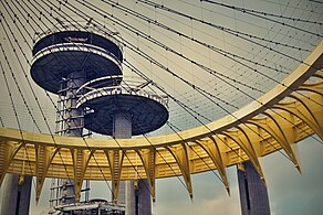 The outer and inner girders of the Tent of Tomorrow, which are curved and painted yellow. There are wires extending inward from the girders. The observation towers are in the background.
