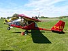 A bright red, high-wing, clear-nosed, pusher-engined aircraft, with yellow registration C-IXII on the tail, sits nose-high in a grassy field with a Piper Cub behind it.