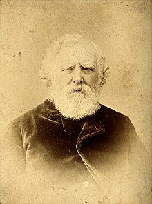 Shown is the very old image of Sir William James Erasmus Wilson in his late years, with white hair and many skin folds. he is wearing a simple siut and wears a full grown beard.