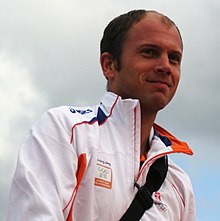 Un homme dégarni portant une veste de sport blanche et orange qui sourit.