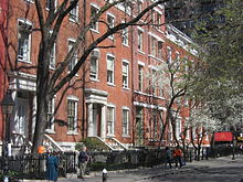 A long shot of a mid-rise apartment building on the side of the street. The building has gates and trees at its front, with trees leaning over the street and pedestrians walking on the sidewalk.