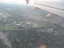 Aerial view of a cloverleaf interchange in a densely popoulated region; an airplane wing is visible at the top of the image.