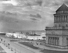 Photographie d'un grand monument de base cubique surmontée par une partie cylindrique à colonnades au sommet de laquelle se trouve un toit conique. Plusieurs cuirassés sont visibles dans le fleuve à l'arrière-plan.