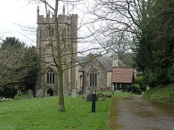 Square gray tower of stone church building, partially obscured by trees. Red roofed lych gate to right. Grass and gravestones in the foreground