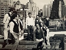 The Stanford Fleet Street Singers perform on the unfinished roof of a medium-height building in New York City, circa 1990. They're dressed in casual outfits with vests and bow ties.