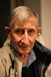 Photographie en couleurs d'un visage masculin vu de face, ayant une large mèche de cheveux gris rejetée sur le front droit.