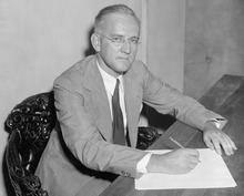 A white man in eyeglasses and a light suit sits at a desk writing a document. He is looking at the camera, possibly posing for this black and white photo.