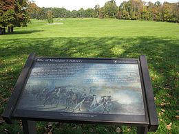 Photo shows an historical sign and a downhill slope toward a road.