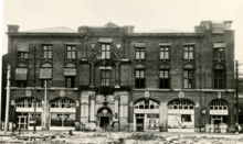 Black and white photo of a three-storey building constructed with dark-coloured bricks, two rows of windows on the upper floors, and larger arched windows on the ground floor