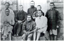 Photographie en noir et blanc de trois prêtres européens en soutane noire portant la barbe, et de cinq jeunes hommes polynésiens.