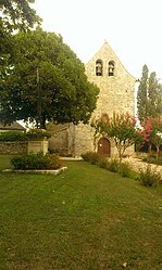 Vue de l'église à une seule nef, en pierre calcaire blanche et toit en tuiles plates, elle n'a pas de clocher individualisé, on voit la place et le calvaire devant l'église