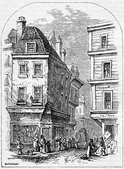 People congregate at the entrance to a narrow street, overlooked by two four-storey buildings. Each floor of the right-most building projects further over the street than the floor below. At the corner of each building, shops advertise their wares. A cart is visible down the street, and one man appears to be carrying a large leg of meat.