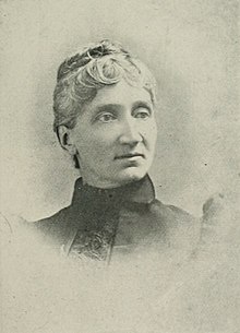 Middle-aged woman with her hair in an up-do wearing a high-neck dark blouse.