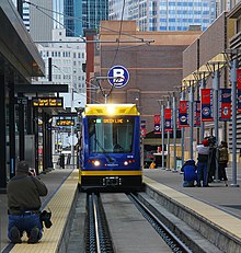 A light rail train with overhead electrification rolls toward the camera in an at-grade rail station.