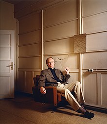 Richard Ford sitting on a couch holding his fist up with a punched-in wall behind him.